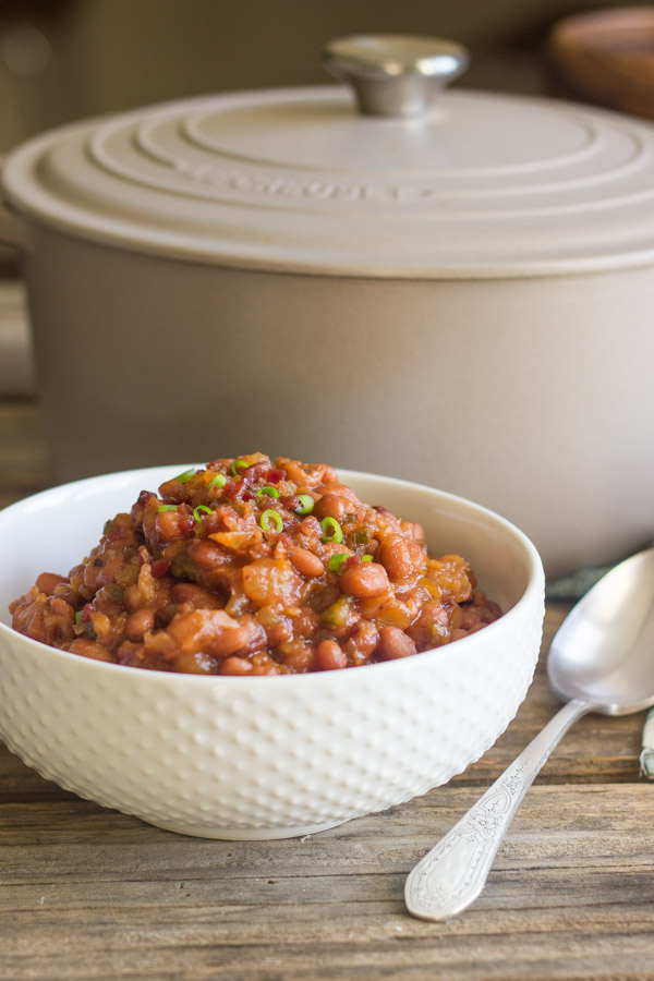 Sweet And Spicy BBQ Baked Beans in a bowl with a spoon next to it and a Le Creuset French Oven in the background.  