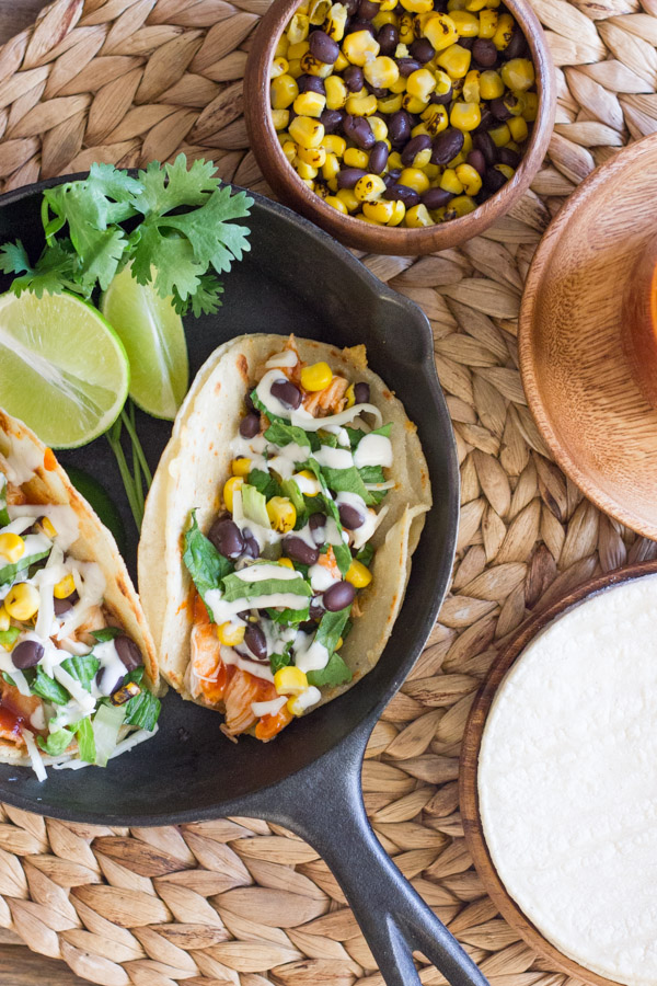 BBQ Chicken Tacos arranged in a mini cast iron skillet with some lime wedges and fresh cilantro, sitting next to a small bowl of corn and black beens, and a plate with tortillas on it.  