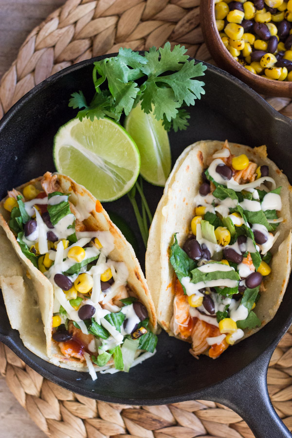 BBQ Chicken Tacos arranged in a mini cast iron skillet with some lime wedges and fresh cilantro, with a small bowl of corn and black beens sitting next to it.  