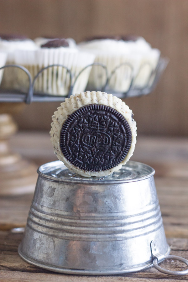 Cookies and Cream Cheesecake Cup sitting on its side showing the Oreo cookie bottom on a mini galvanized bucket that is upside down, with more Cookies and Cream Cheesecake Cups arranged on a cake stand in the background.  