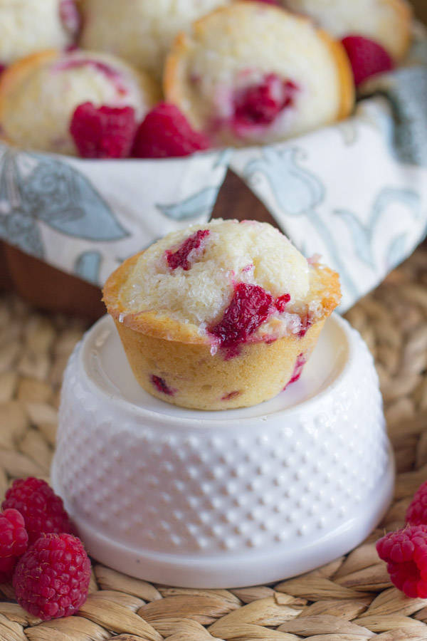 Healthier Raspberry Almond Muffin sitting on top of a glass dish with fresh raspberries around it, and a cloth lined bowl full of muffins in the background.  