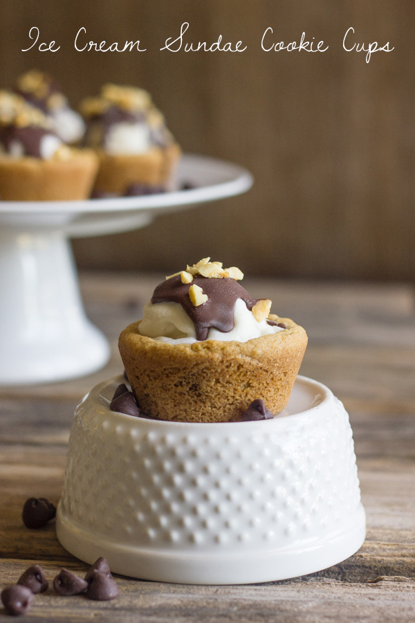 Ice Cream Sundae Cookie Cup sitting on top of a glass bowl, with chocolate chips around it, and a cake stand of more Ice Cream Sundae Cookie Cups in the background. 