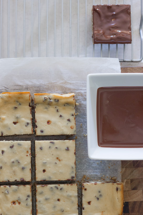 Oatmeal Chocolate Chip Cheesecake Bars sitting on parchment paper next to a square bowl of melted chocolate, with a dipped Oatmeal Chocolate Chip Cheesecake Bar on a cooling rack.  