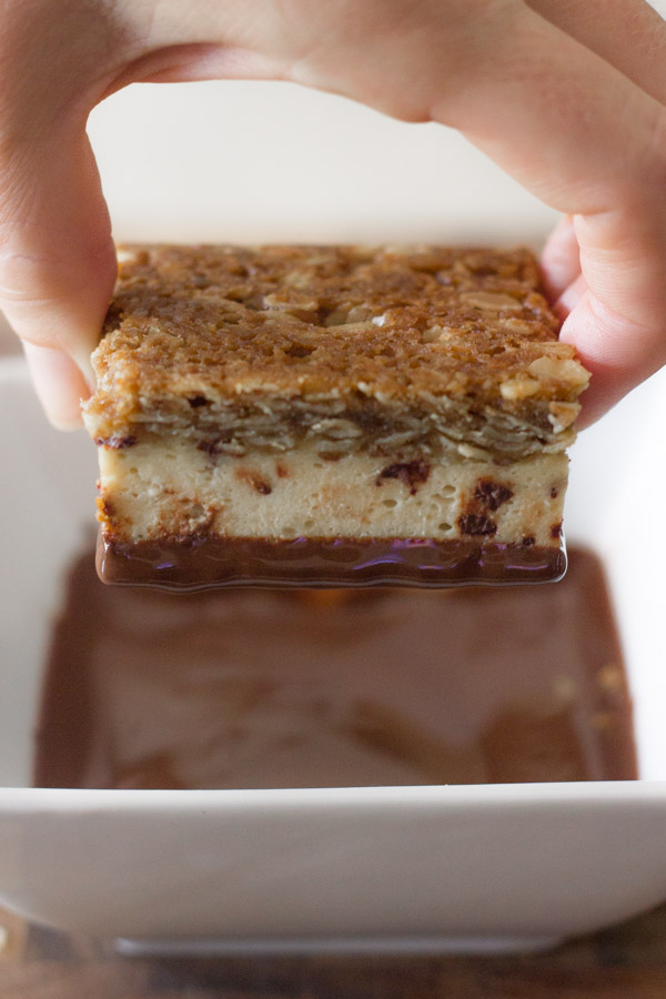 Oatmeal Chocolate Chip Cheesecake Bar being held over a square bowl of melted chocolate. 