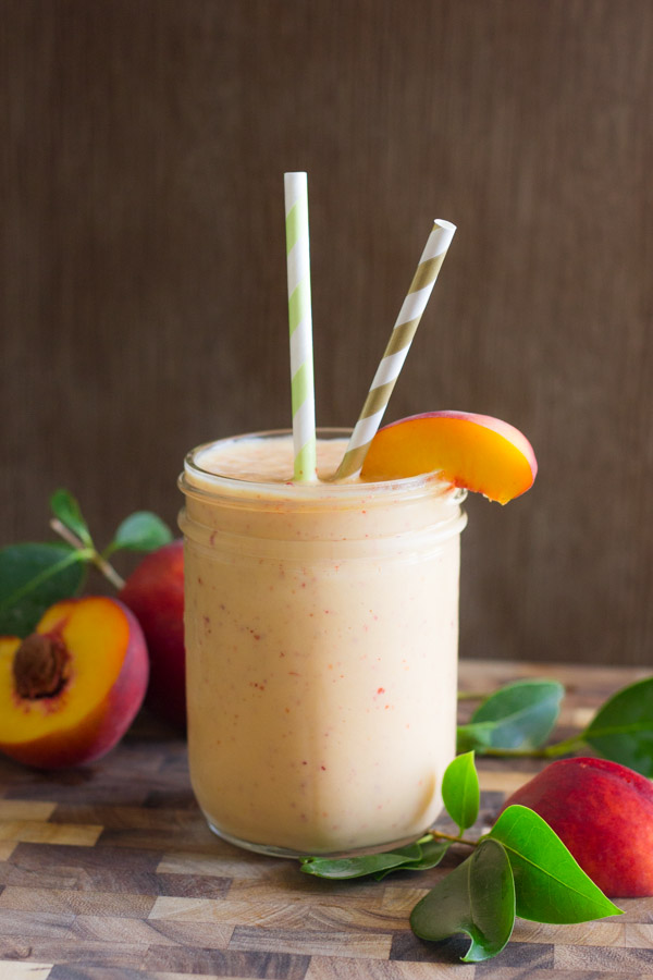 Skinny Peach Cream Slush in a glass with two straws and a peach slice, sitting on a cutting board with peaches in the background.  