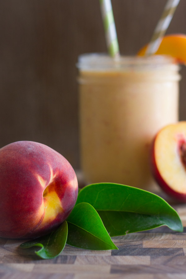 A whole peach on a cutting board, with a Skinny Peach Cream Slush in a glass with two straws and a peach slice in the background.  