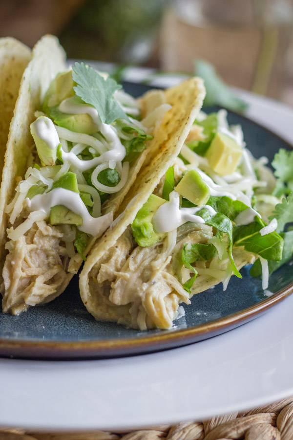 Easy Creamy Crockpot Salsa Verde Chicken in crunchy taco shells with shredded lettuce, diced avocado, sour cream and cilantro, on a plate.  