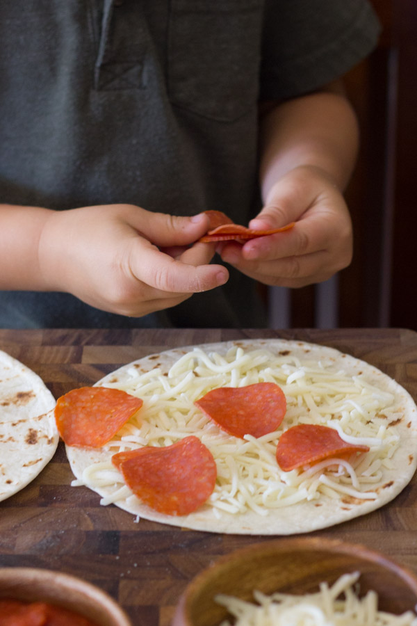 A tortilla with cheese and pepperoni on it, with more pepperoni being placed on it by a child.  