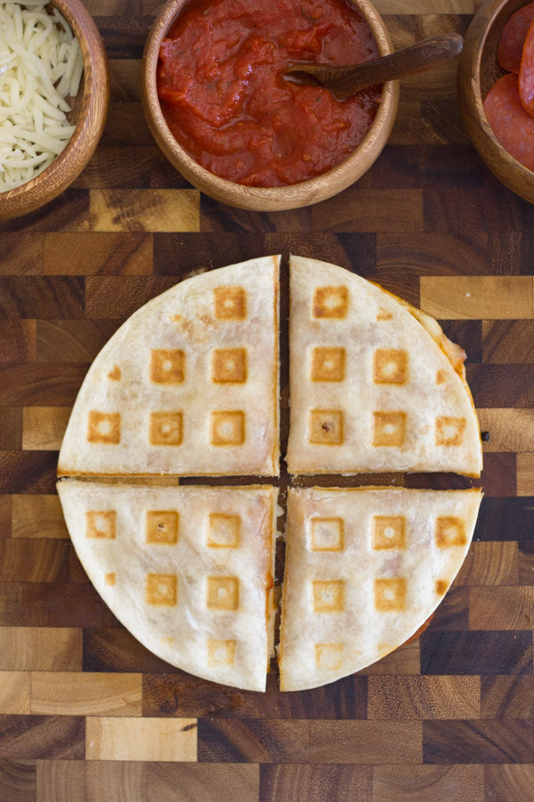 Waffled Pizza Pocket cut in four pieces on at cutting board with a bowl of mozzarella cheese, a bowl of pizza sauce and a bowl of pepperoni next to it.  