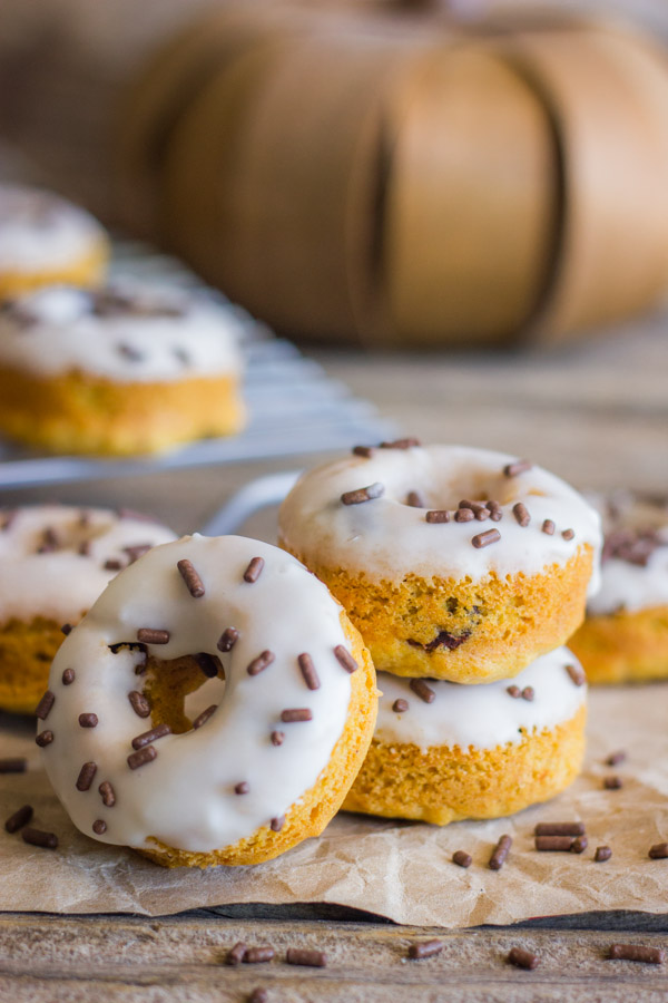 Mini Baked Pumpkin Chocolate Chip Donuts stacked on paper with sprinkles around them, with more Mini Baked Pumpkin Chocolate Chip Donuts on a cooling rack in the background with a pumpkin decoration.  