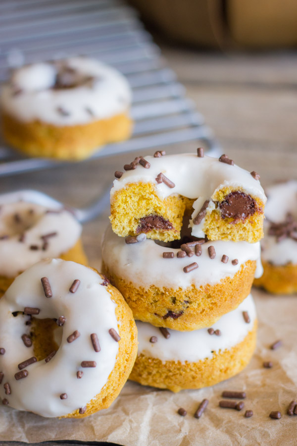 Mini Baked Pumpkin Chocolate Chip Donuts stacked on paper with sprinkles around them and the donut on top is cut in half, with more Mini Baked Pumpkin Chocolate Chip Donuts on a cooling rack in the background.