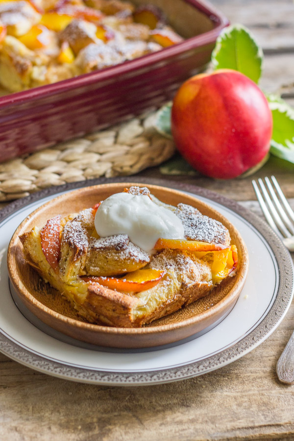 Peaches and Cream French Toast Bake topped homemade whipped cream on a plate, with the baking dish of Peaches and Cream French Toast in the background with a whole peach.  