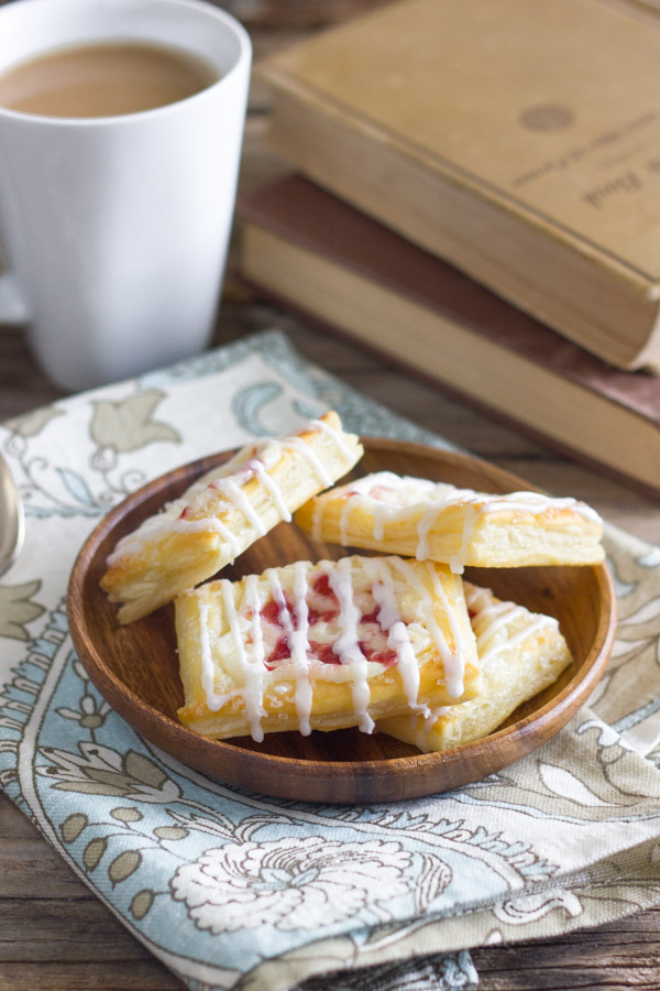Raspberry Cream Cheese Danishes on a wood plate that is sitting on a cloth napkin, with a cup of coffee and stacked books in the background.  