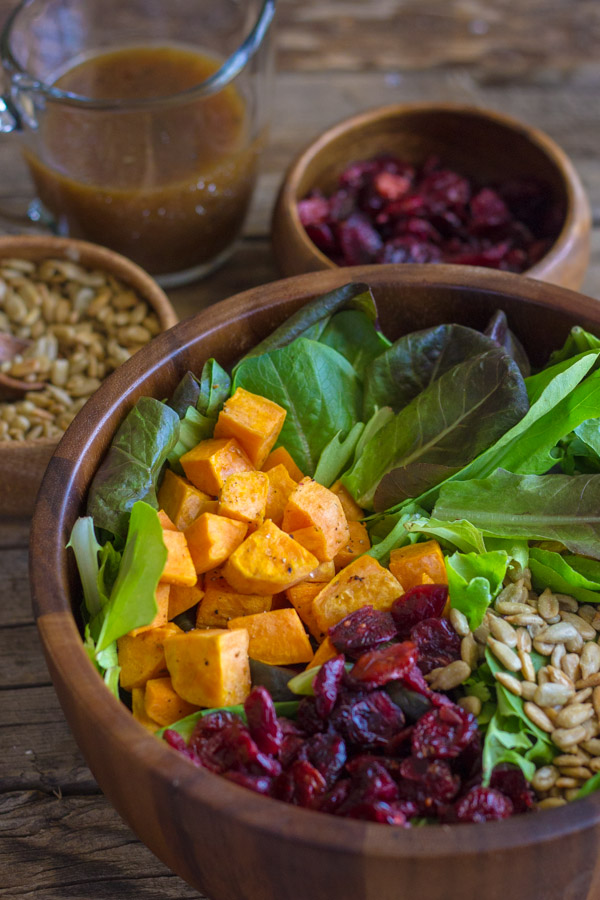 Roasted Sweet Potato Salad ingredients in a wood bowl, with two small bowls of sunflower seeds and dried cranberries, along with a small glass pitcher of dressing in the background.  