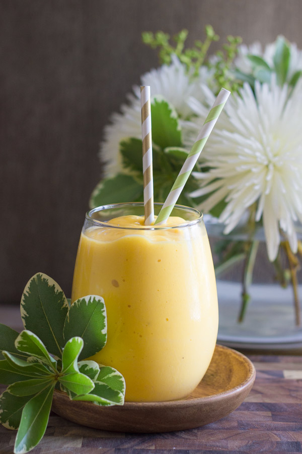 Tropical Sunshine Smoothie in a glass with two straws sitting on a little wood saucer, with a flower arrangement in the background.  