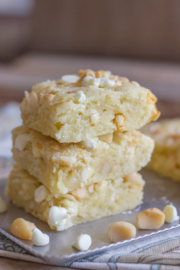 White Chocolate Macadamia Nut Bars stacked in a pile of three on a serving platter, with a few macadamia nuts and white chocolate chips around them.  
