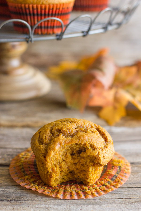 Best Ever Pumpkin Muffin with its wrapper pulled down and a bite taken out of it, with more muffins on a metal cake stand in the background.  