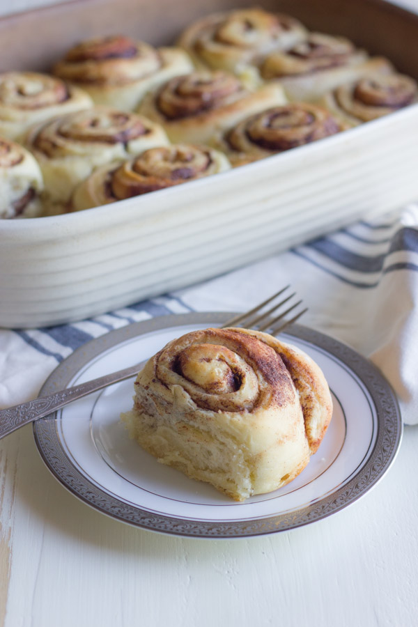 Overnight Cinnamon Roll on a plate with a fork, with a baking dish of Overnight Cinnamon Rolls in the background.  