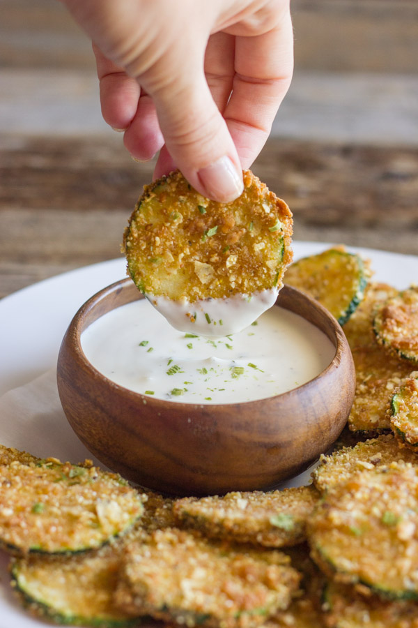 Pita Chip Fried Zucchini on a serving plate with a small bowl of ranch dressing, and one Pita Chip Fried Zucchini slice being dipped into the ranch dressing.  