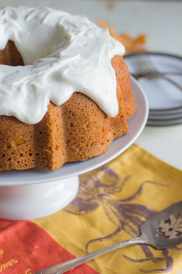 Pumpkin Spice Bundt Cake with frosting on a cake stand, with a serving utensil and a plate with forks next to the stand.  