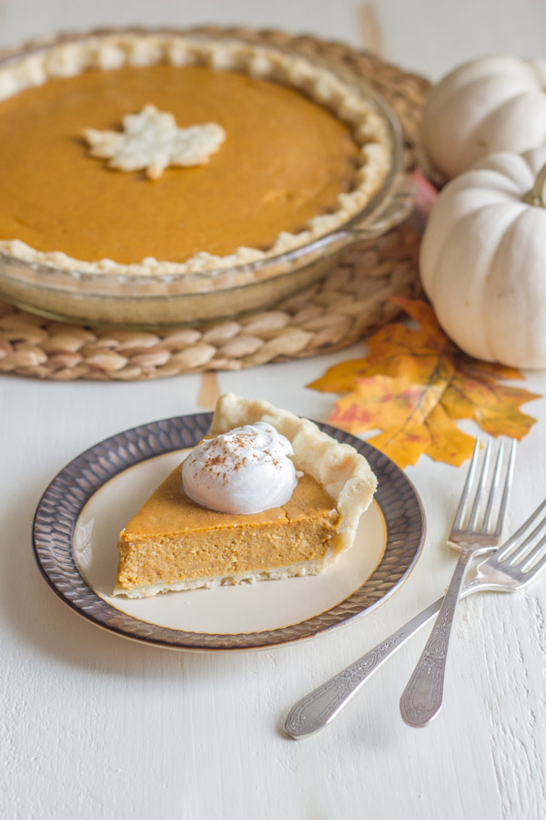 Classic Pumpkin Pie piece on a plate topped with whipped cream, with a whole Classic Pumpkin Pie in the background. 