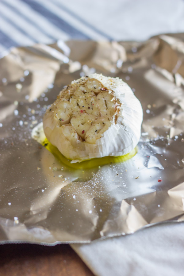 A head of garlic with the top sliced off, that has been drizzled with olive oil and sprinkled salt and pepper, sitting on a piece of tin foil.