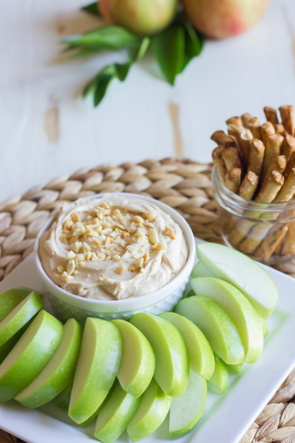 Greek Yogurt Peanut Butter Dip in a bowl, sitting on a serving platter with sliced apples, and a glass jar of stick pretzels next to it.  