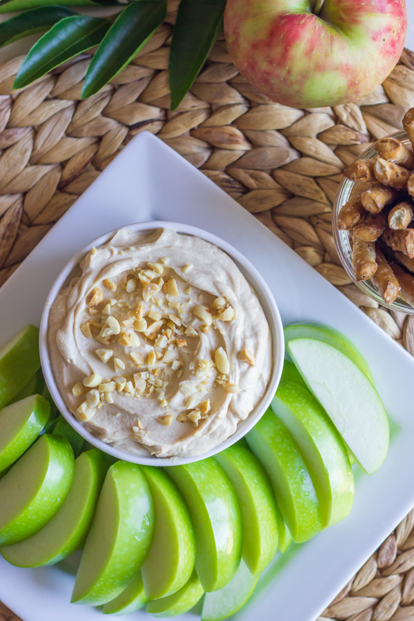 Greek Yogurt Peanut Butter Dip in a bowl, sitting on a serving platter with sliced apples, and a glass jar of stick pretzels next to it.  