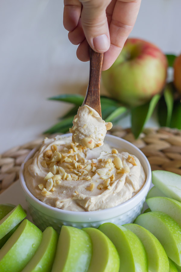 Greek Yogurt Peanut Butter Dip in a bowl, sitting on a serving platter with sliced apples, and a tiny spoon with dip on it.  