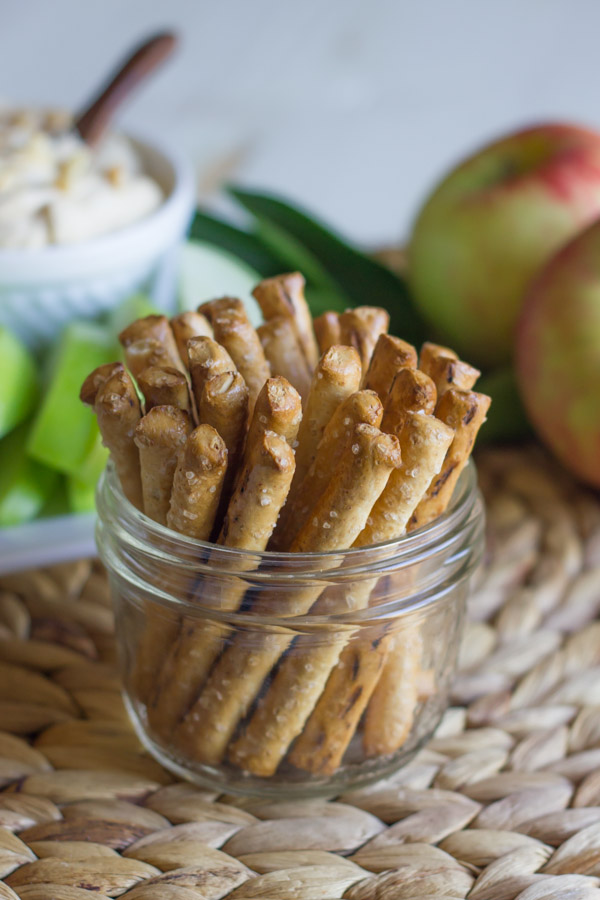A glass jar of stick pretzels, with Greek Yogurt Peanut Butter Dip in a bowl sitting on a serving platter with sliced apples in the background, along with some whole apples.   