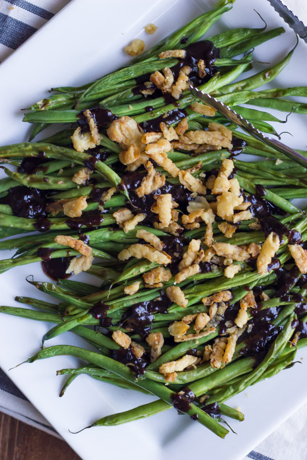 Roasted Green Beans with Creamy Cranberry Balsamic topped with french friend onions on a serving plate with tongs. 