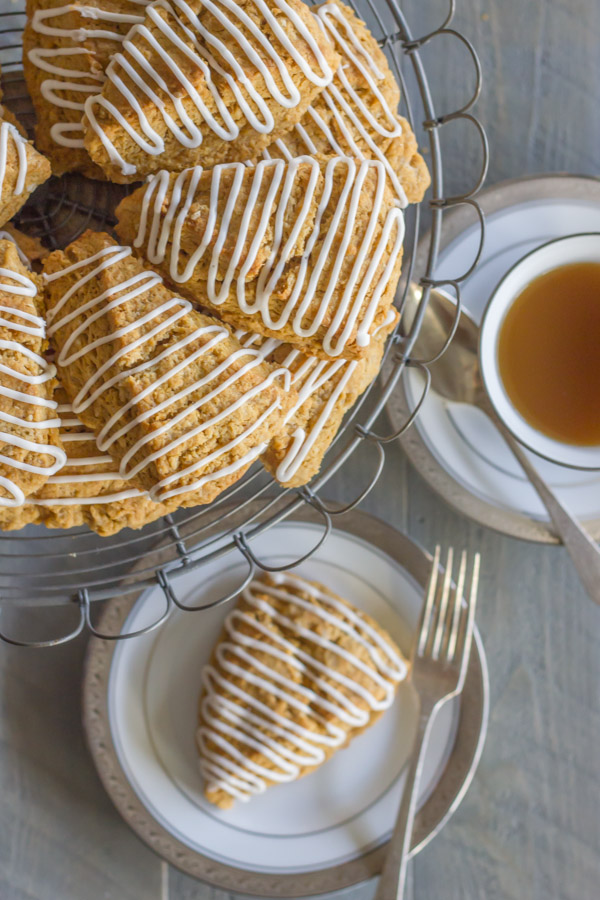 Iced Gingerbread Oat Scone on a plate with a fork, and more scones on a cake stand next to it, as well as a cup of coffee.  