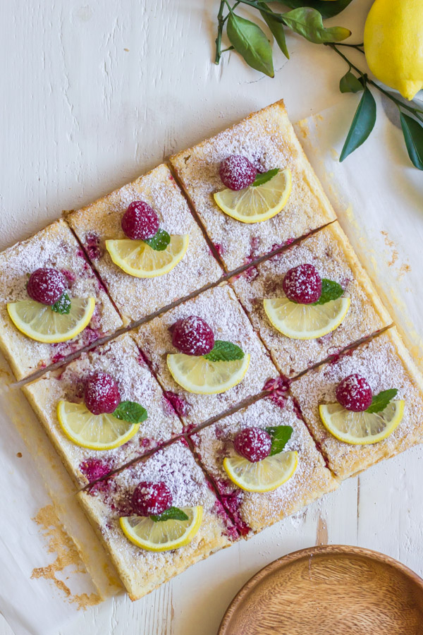 Raspberry Lemon Bars cut into squares, each one garnished with a lemon slice, a whole raspberry and a mint leaf.  