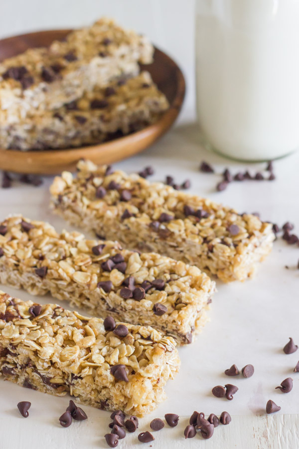Three Copycat Quaker Chewy Chocolate Chip Granola Bars next to each other, with a glass of milk and more bars on a plate in the background, and chocolate chips sprinkled around.   