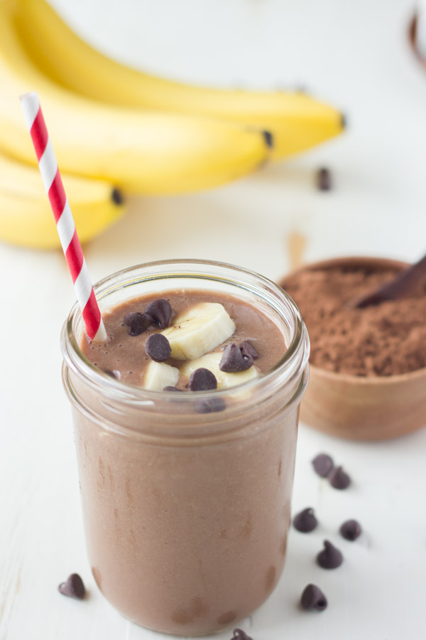 Chocolate Banana Smoothie topped with banana slices and chocolate chips, in a glass jar with a straw, with a small bowl of cocoa powder and a bunch of whole bananas in the background. 