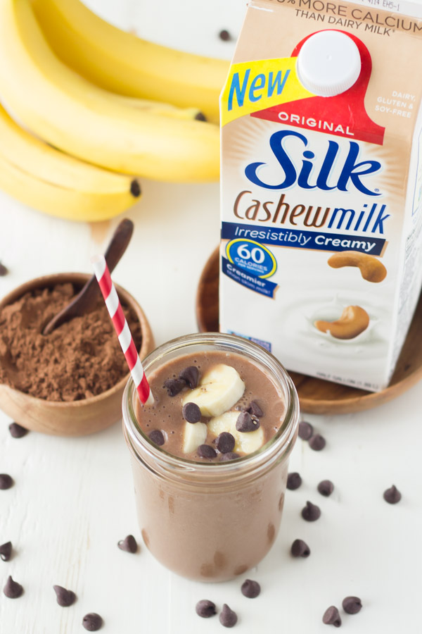 Chocolate Banana Smoothie topped with banana slices and chocolate chips, in a glass jar with a straw, sitting next to a small bowl of cocoa powder, a bunch of whole bananas and a carton of Silk Cashewmilk. 