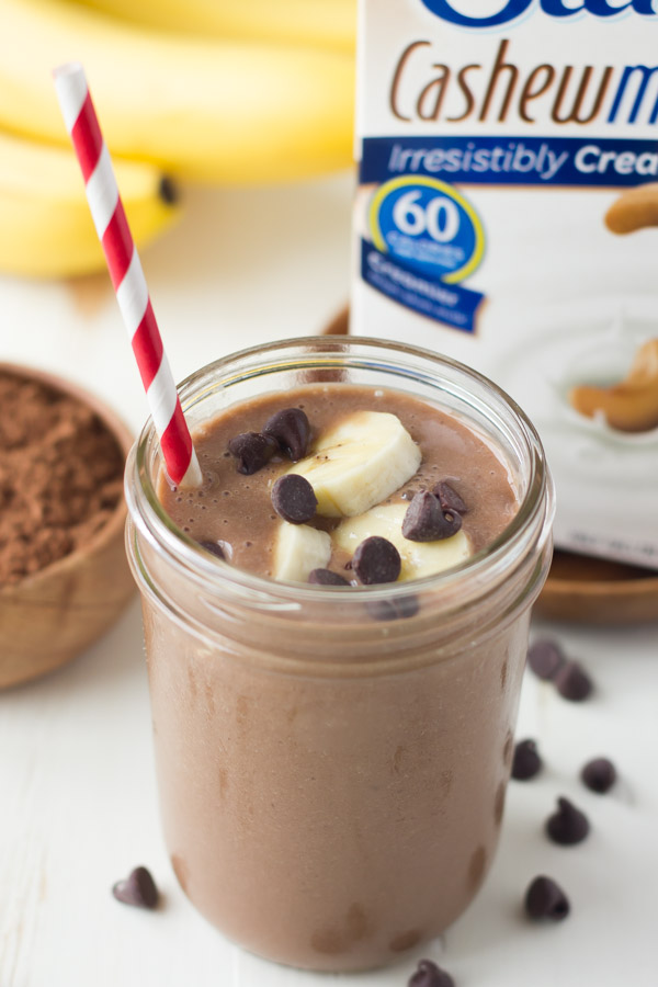 Chocolate Banana Smoothie topped with banana slices and chocolate chips, in a glass jar with a straw, with a small bowl of cocoa powder, a bunch of whole bananas and a carton of Silk Cashewmilk in the background. 