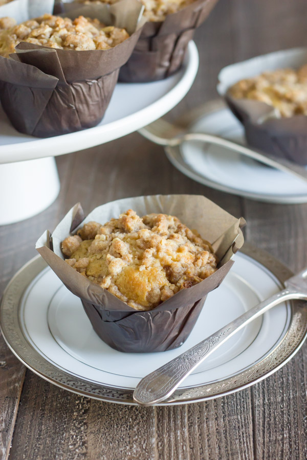 Healthier Cinnamon Oat Streusel Muffin in a parchment paper muffin liner on a plate, with more muffins on a cake stand in the background along with another muffin on a plate.  