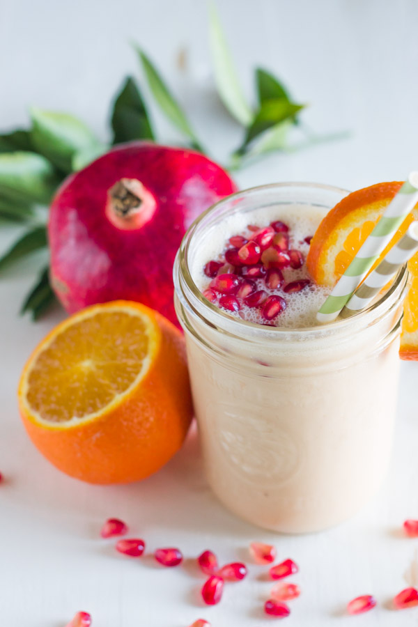 Pomegranate Citrus Smoothie in a glass jar with pomegranate seeds on top, an orange slice on the rim of the glass and two straws in it, with an orange and pomegranate next to the glass.  