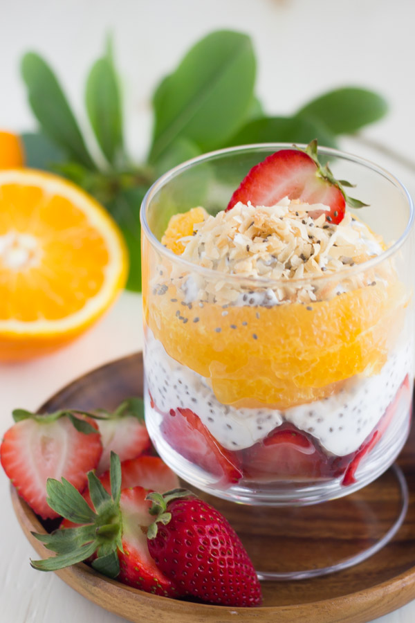 Toasted Coconut Chia Yogurt Fruit Parfait in a glass cup, sitting on a wood plate with sliced strawberries.  
