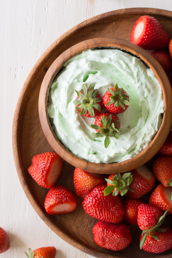 Creamy Pistachio Fruit Dip in a wood bowl topped with three whole strawberries, sitting on a wood plate with more whole strawberries. 