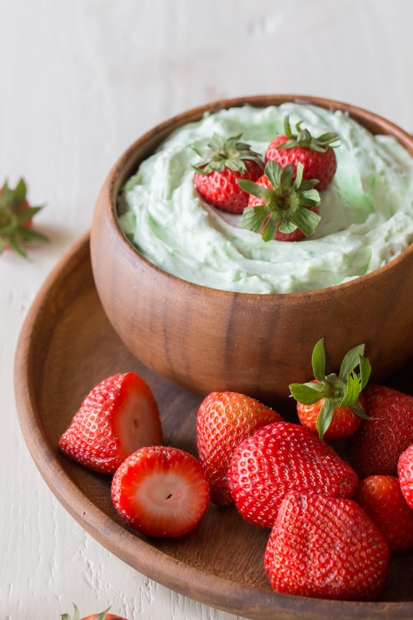 Creamy Pistachio Fruit Dip in a wood bowl topped with three whole strawberries, sitting on a wood plate with more whole strawberries. 