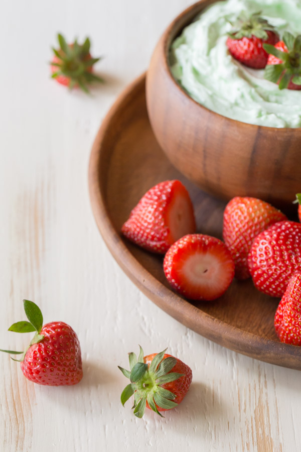 Creamy Pistachio Fruit Dip in a wood bowl topped with a few whole strawberries, sitting on a wood plate with more whole strawberries. 
