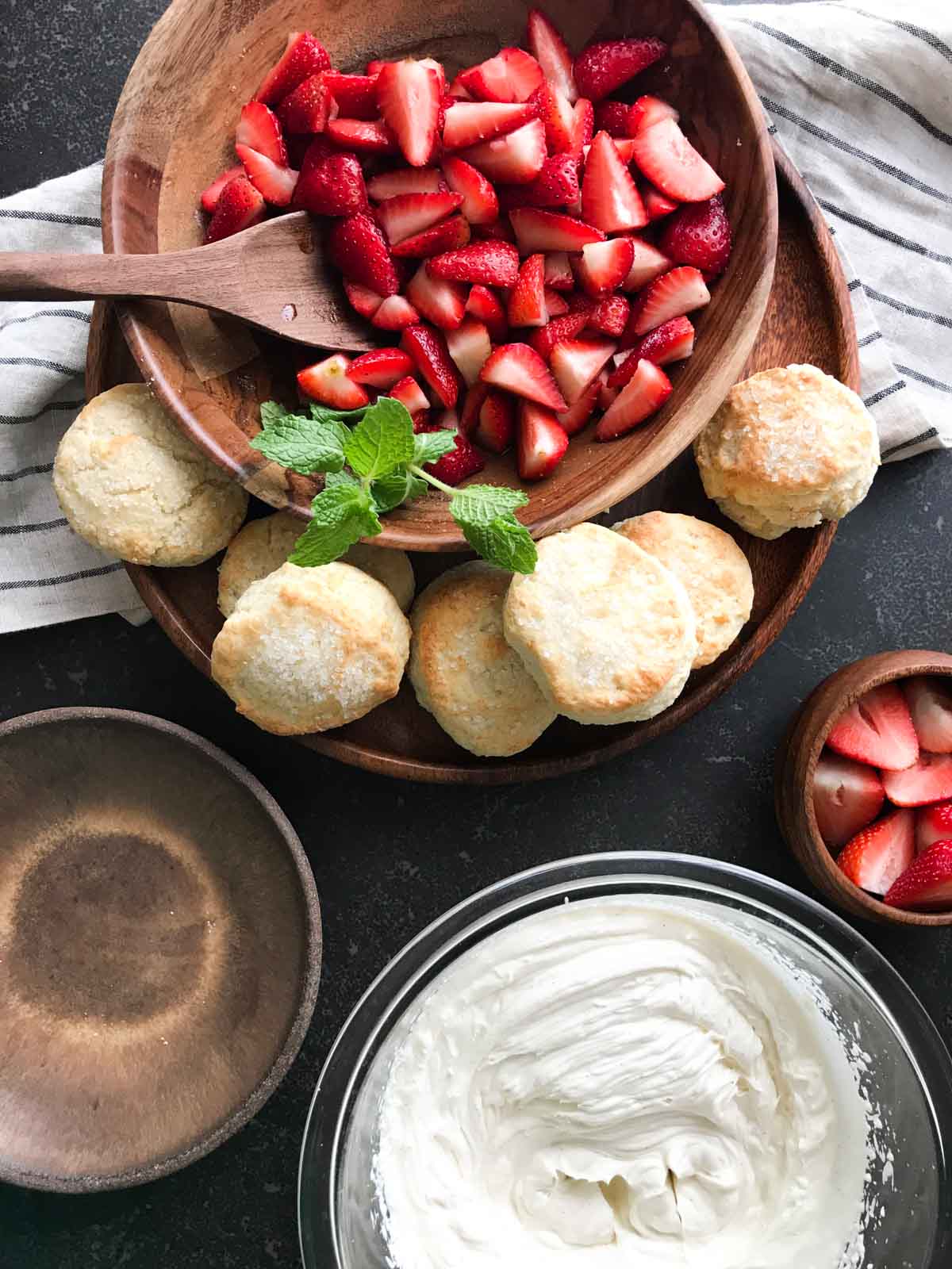 All the ingredients for the Homemade Strawberry Shortcakes ready to be assembled - a bowl of strawberries sitting on a plate with buttermilk biscuits and a bowl of whipped cream. 