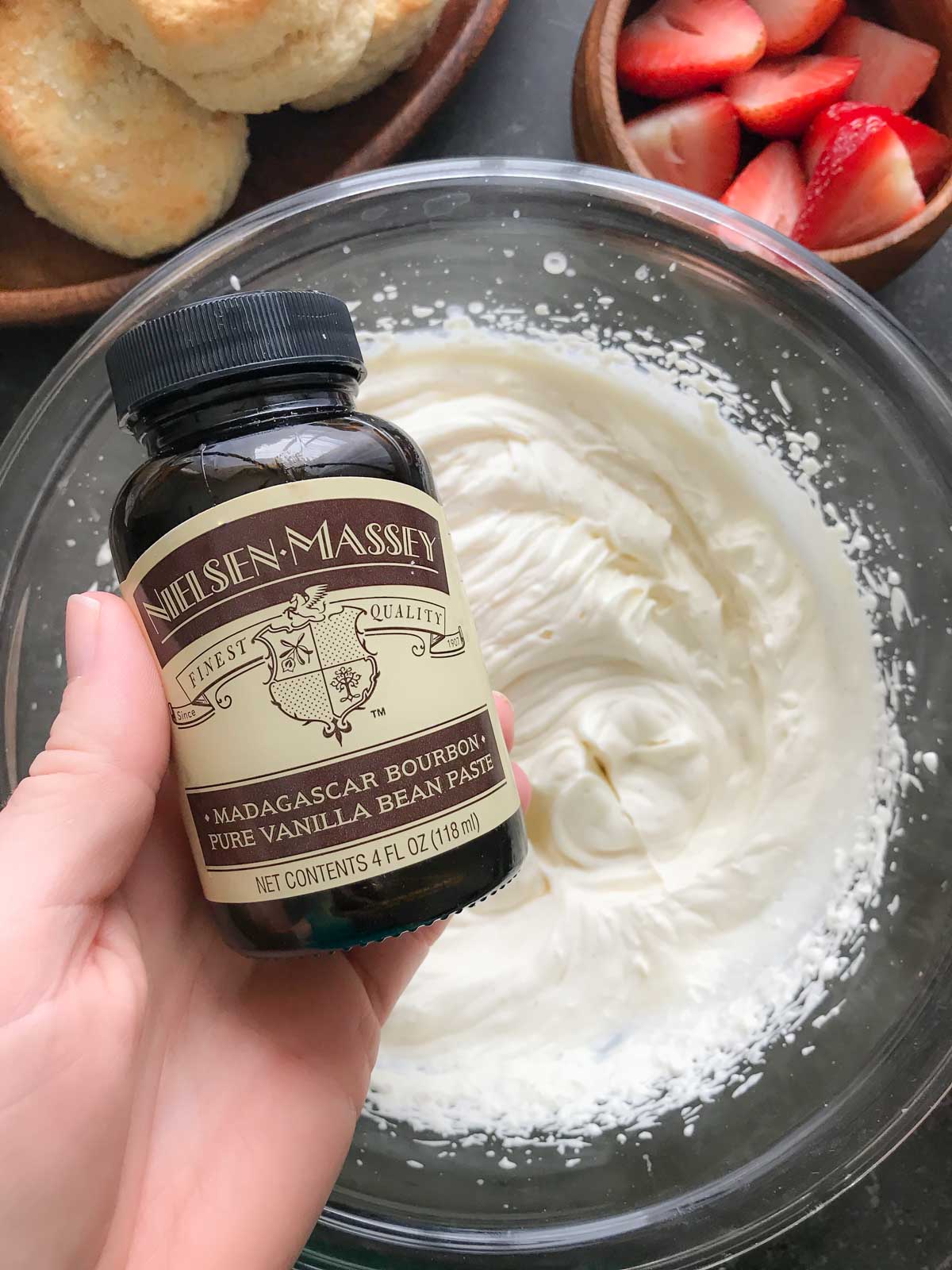 A jar of Madagascar Bourbon Vanilla Bean Paste from Nielsen Massey held over a bowl of homemade whipped cream. 