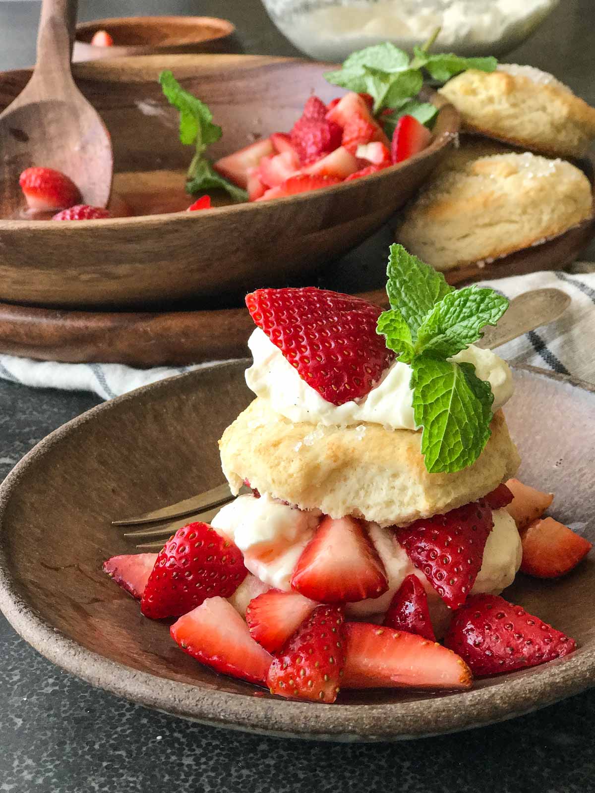 A Homemade Strawberry Shortcake assembled in a bowl with a fork, with the ingredients for the Strawberry Shortcakes in the background. 