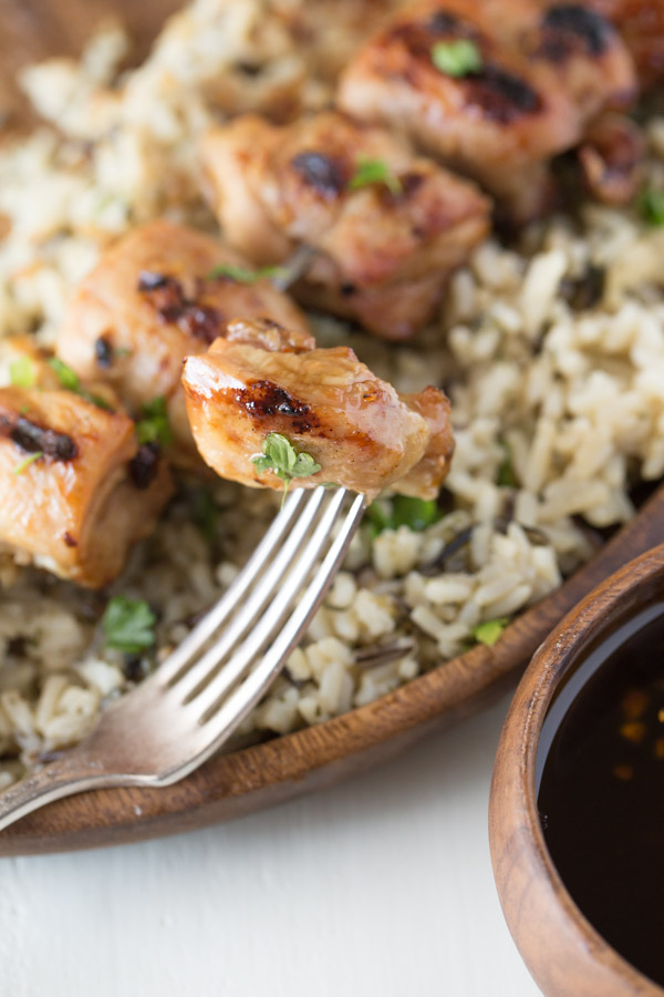 A piece of Maple Dijon Glazed Chicken on a fork, with Maple Dijon Glazed Chicken served over a bed of rice on a wood plate in the background, along with a small bowl of the Maple Dijon Glaze. 