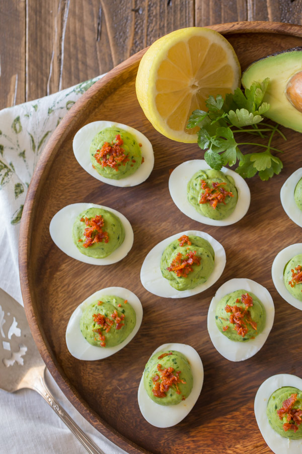 Smoky Bacon Avocado Deviled Eggs arranged on a wood serving plate, with a half of an avocado, a half of a lemon and a little bunch of cilantro.  
