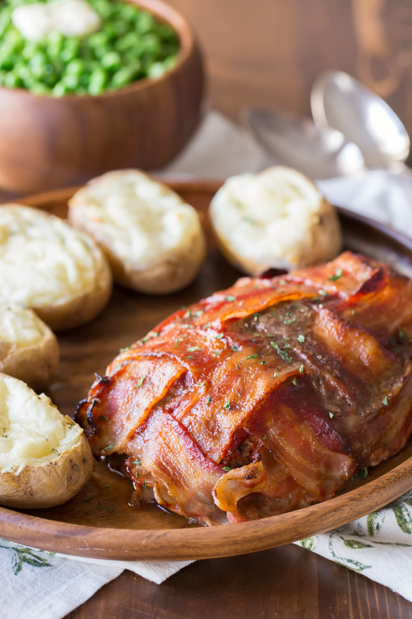 Bacon Wrapped Meatloaf on a wood serving plate with five twice baked potatoes, and a bowl of peas in the background.  