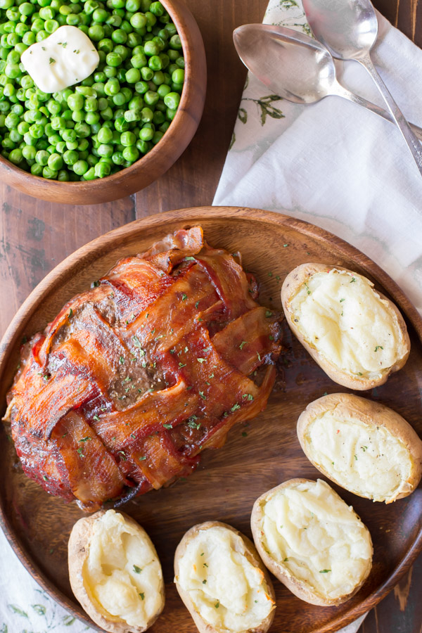 Bacon Wrapped Meatloaf on a wood serving plate with five twice baked potatoes, and a bowl of peas next to it.  